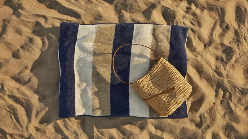 Grosses gestreiftes Strandtuch auf warmem Sand neben einer geflochtenen Strohtasche, von oben gesehen